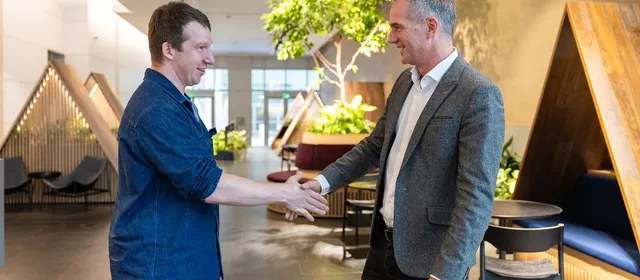 Two men shake hands in a bright office lobby with wooden booth seating and an indoor tree.