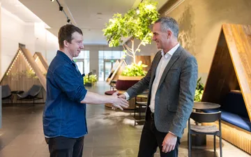 Two men shake hands in a bright office lobby with wooden booth seating and an indoor tree.