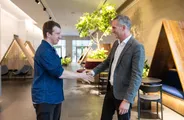 Two men shake hands in a bright office lobby with wooden booth seating and an indoor tree.